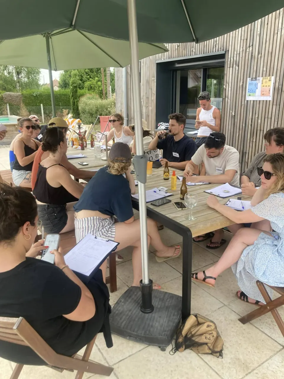 Tablée de joueurs en terrasse sous un parasol, carnets d'enquête en main